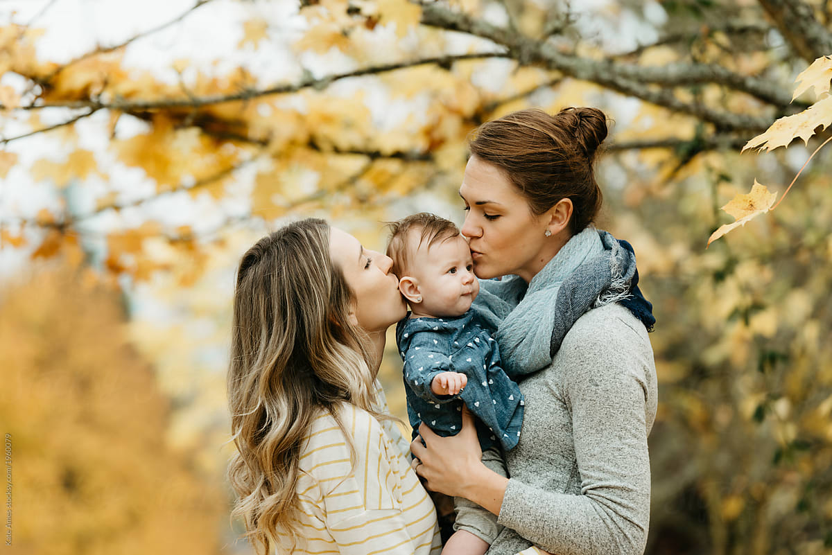 Two smiling moms walking with their baby daughter in a park, symbolizing LGBTQIA+ co-parenting and rainbow family inclusion.