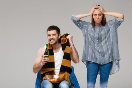Frustrated woman holding her head next to an angry man with a beer bottle, wearing a sports scarf and holding a bowl of chips—illustrating emotional tension and unhealthy behavior in a co-parenting dynamic.