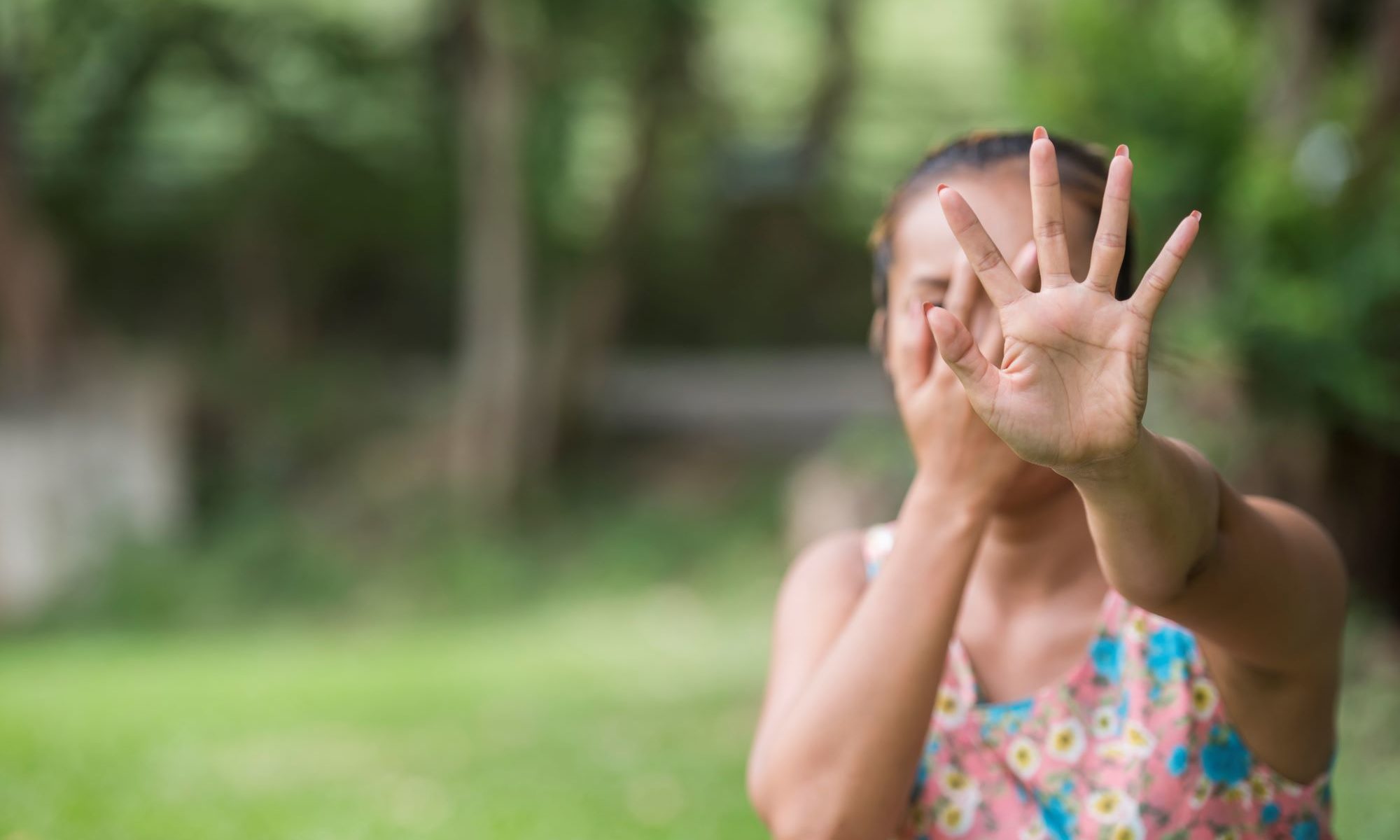 A woman in a floral dress holds up her hand toward the camera in a clear “stop” gesture while covering part of her face, expressing fear or distress. The blurred natural background contrasts with her emotional state—highlighting the urgency of protecting oneself from an ex-partner’s harassment and establishing safe, respectful co-parenting boundaries.