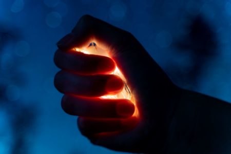 Close-up of a hand gripping a strand of glowing lights in a dark blue background, evoking a sense of emotional intensity and restraint. The image reflects the inner pressure a child may feel when caught between co-parents and the importance of helping them release that tension—“ungashlisht”—through empathy, structure, and safe expression.