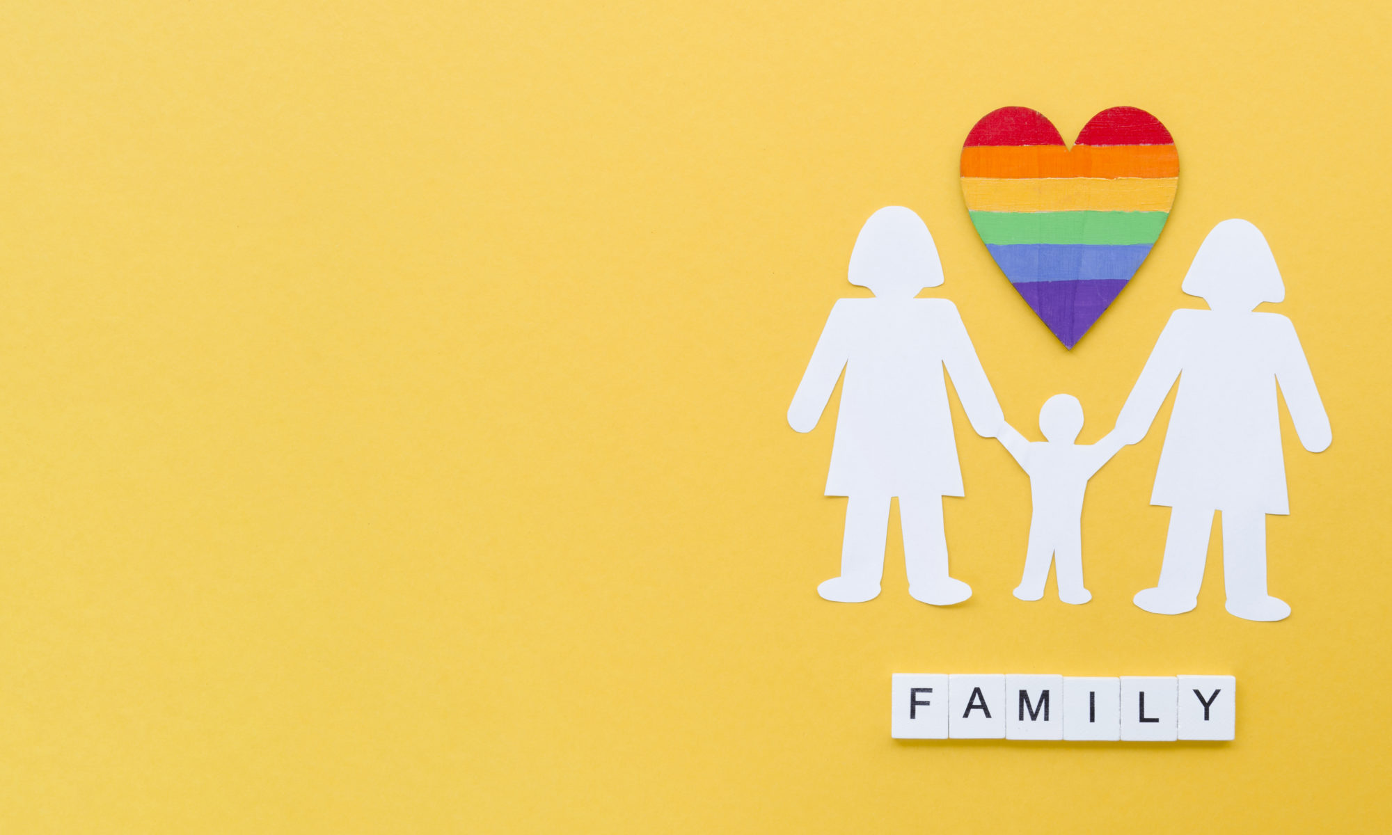 Illustration of an LGBTQ+ family with two female parents and a child holding hands under a rainbow heart, symbolizing love, diversity, and co-parenting. The word 'FAMILY' is spelled out beneath them on a yellow background.