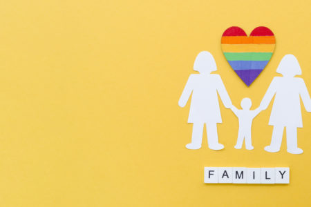 Illustration of an LGBTQ+ family with two female parents and a child holding hands under a rainbow heart, symbolizing love, diversity, and co-parenting. The word 'FAMILY' is spelled out beneath them on a yellow background.
