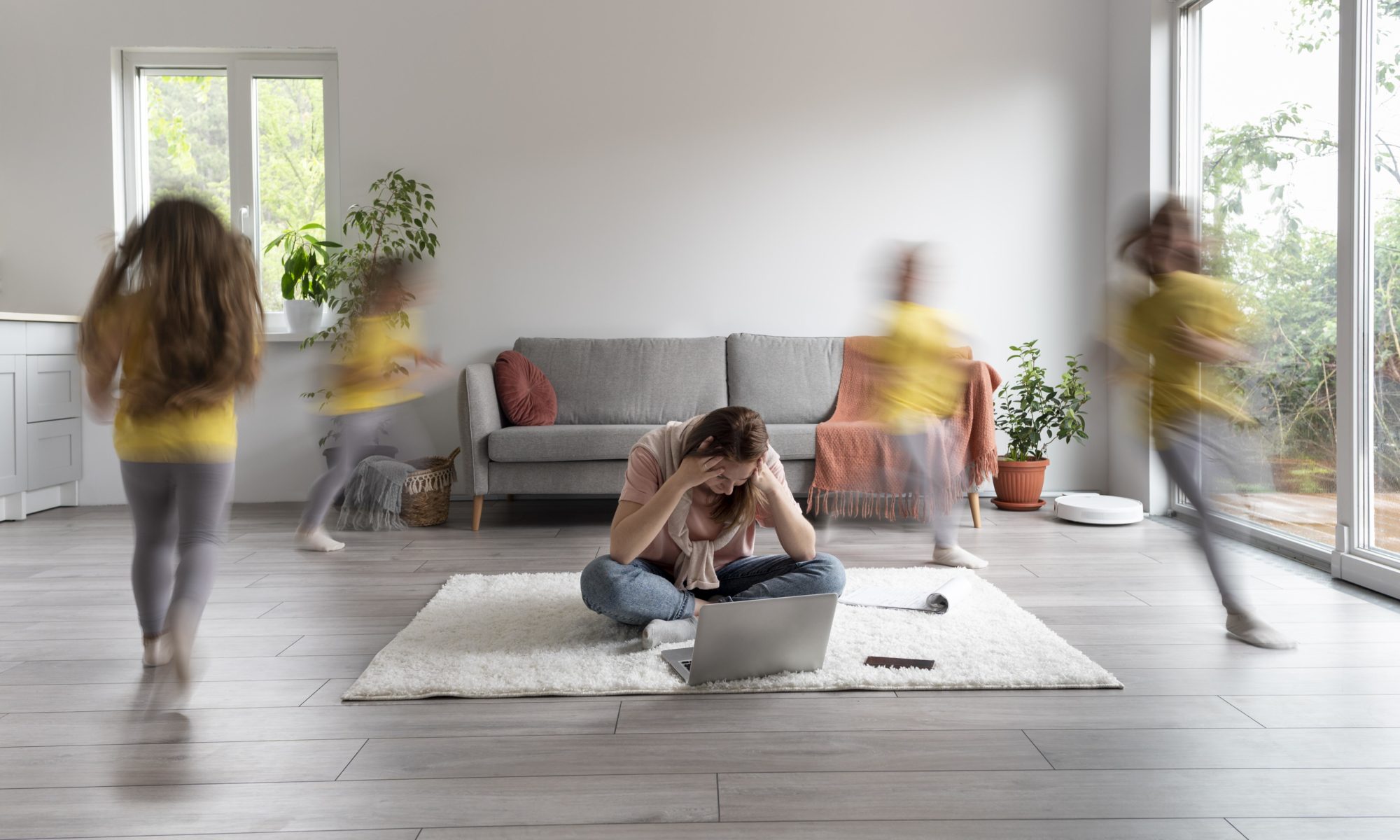 Stressed mother sitting on the floor with a laptop, surrounded by blurred children running around — a visual representation of parental burnout and emotional overwhelm in modern parenting.