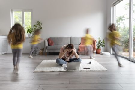 Stressed mother sitting on the floor with a laptop, surrounded by blurred children running around — a visual representation of parental burnout and emotional overwhelm in modern parenting.