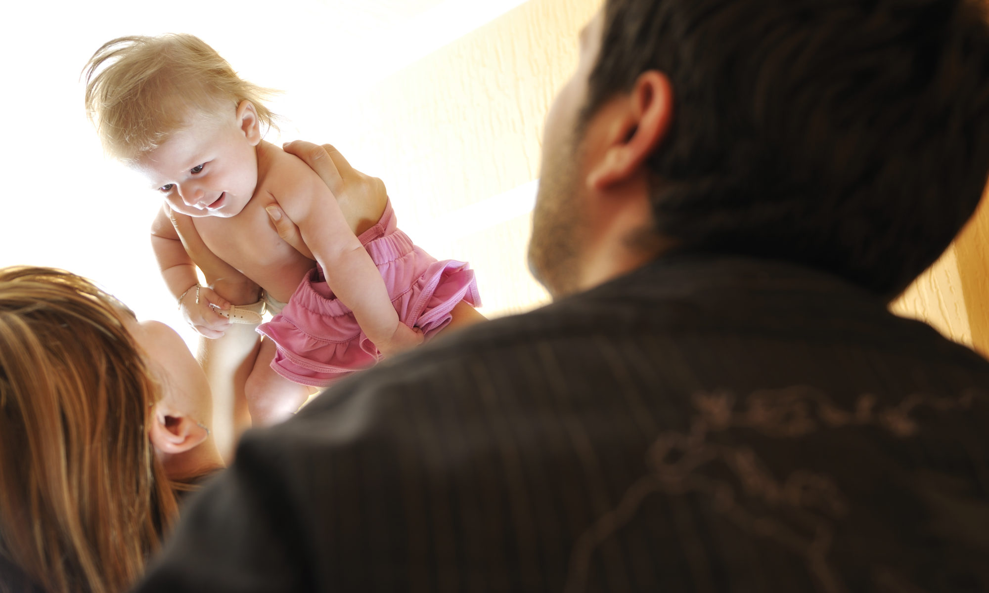 A smiling baby in a pink skirt is lifted into the air by one adult while another watches affectionately. The scene is filled with warm light, capturing a joyful and caring moment between parents and child in a supervised parenting context.