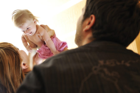 A smiling baby in a pink skirt is lifted into the air by one adult while another watches affectionately. The scene is filled with warm light, capturing a joyful and caring moment between parents and child in a supervised parenting context.